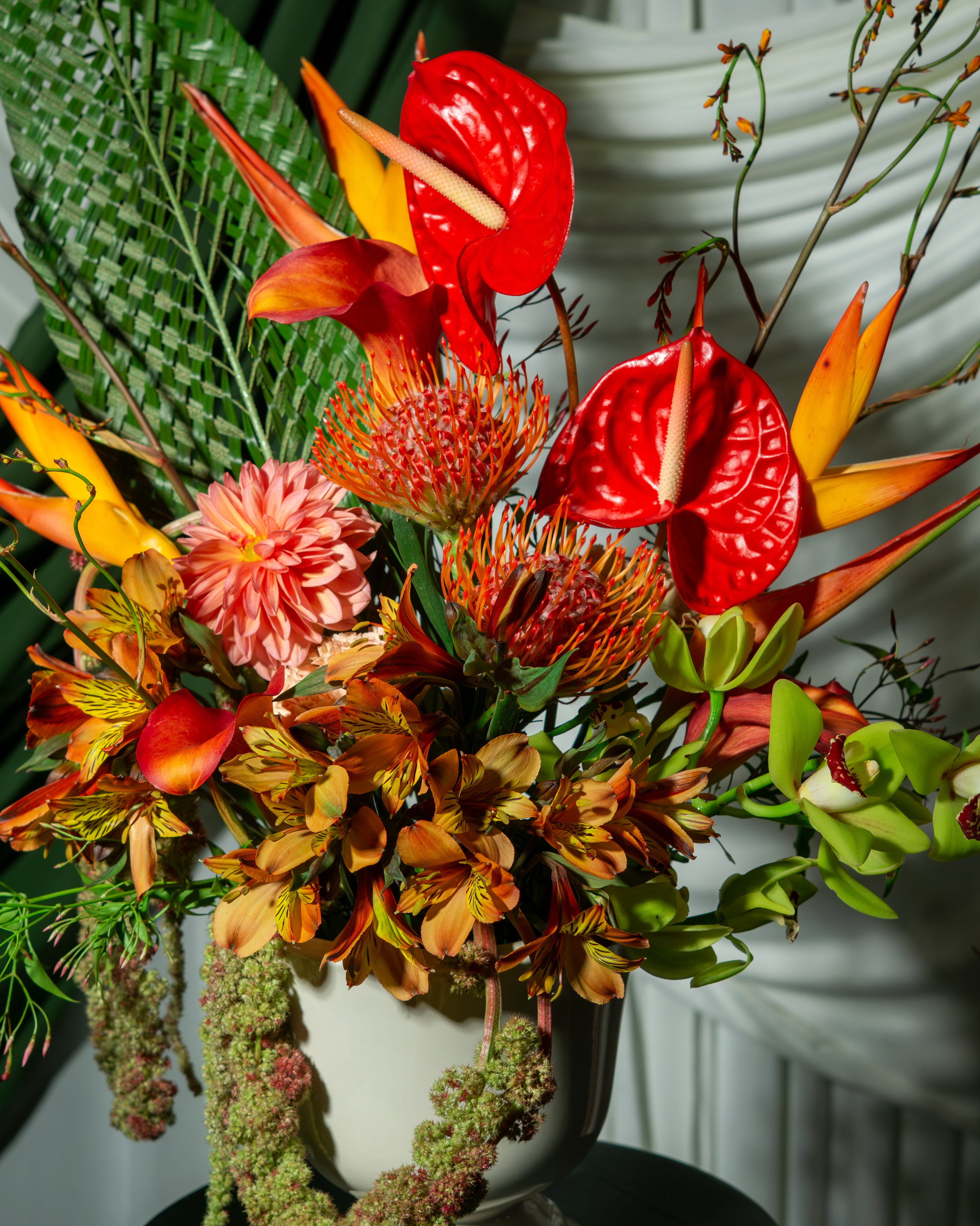 Colorful floral arrangement with red anthuriums, green leaves, and other tropical flowers in a white vase.