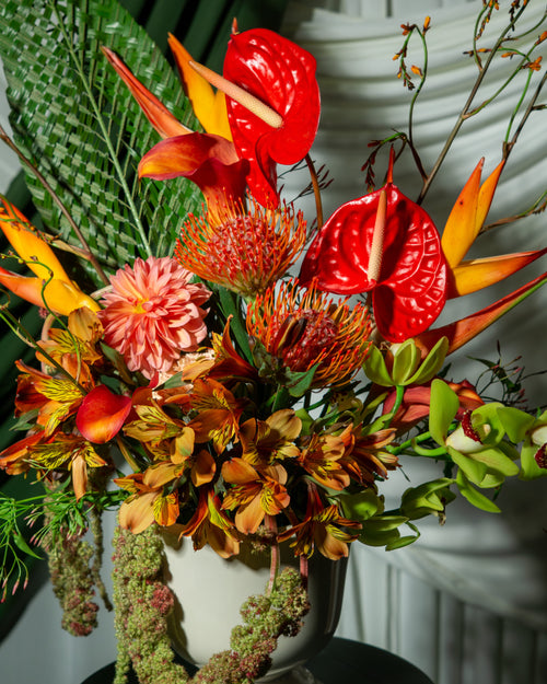 Colorful floral arrangement with red anthuriums, green leaves, and other tropical flowers in a white vase.